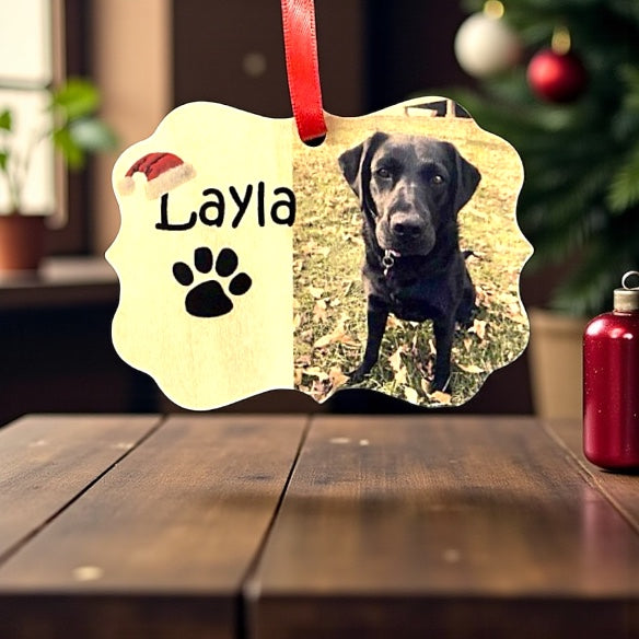 Personalized pet ornament with a black dog named Layla on a wooden surface with a blurred Christmas tree background.