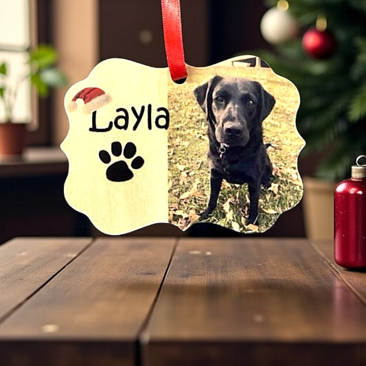 Personalized pet ornament with a black dog named Layla on a wooden surface with a blurred Christmas tree background.